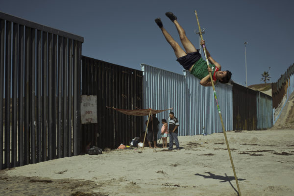 A mexican pole vault jumper trains by the wall in Tijuana beach while a family uses the wall to mount their beach day tent.